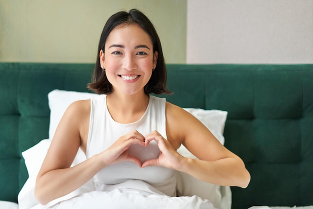 Smiling korean girl shows heart love sign spending time in\
bedroom lying in bed in morning covered w