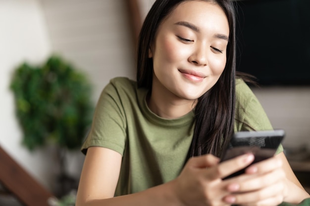 Smiling korean woman looking at mobile phone chat app with dreamy face sitting at home on couch