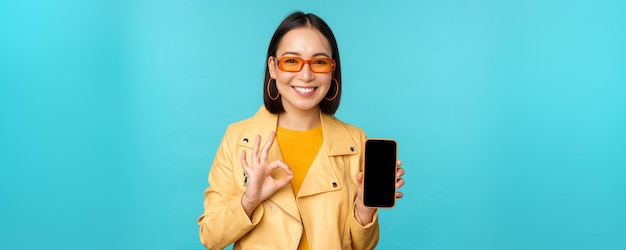 Smiling korean woman showing mobile phone app interface smartphone application recommending on cellphone standing over blue background