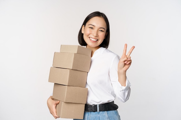 Smiling korean woman with boxes showing vsign peace gesture standing over white background