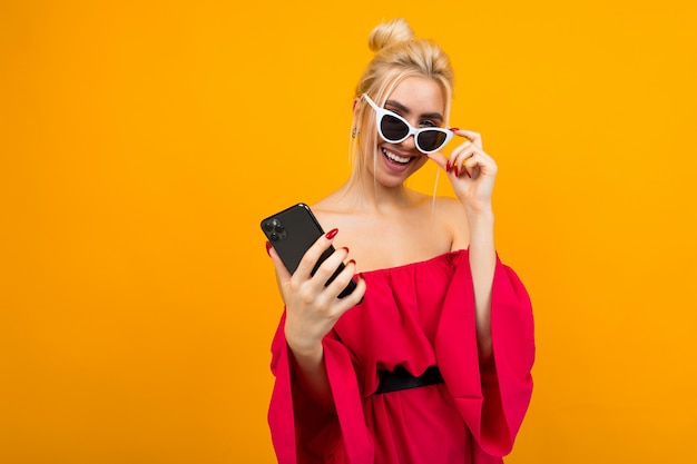 smiling lady in a red dress holds glasses on her face while holding a phone on a yellow surface with copy space