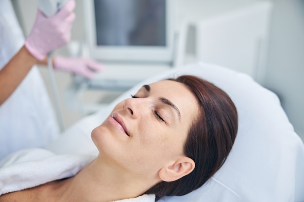 Smiling lady relaxing with her eyes closed before the ultrasound treatment in a cosmetologist office