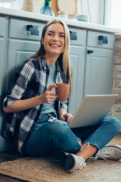 Smiling lady sitting on the floor with a laptop and holding a mug while looking away