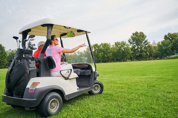 Smiling lady in sunglasses pointing at something to a golf car driver in a cap