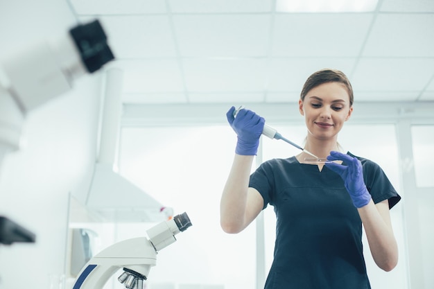 Smiling lady wearing rubber gloves while using an automatic pipette dispenser with a microscope slide