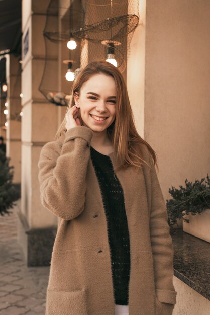 La signora sorridente indossa un cappotto marrone, pone sulla macchina fotografica vicino alla parete marrone del ristorante, guarda la macchina fotografica e sorride francamente