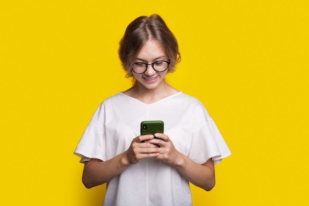 Smiling lady with glasses and blonde hair is chatting on mobile posing on a yellow studio wall