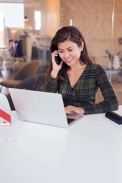 Photo smiling lady working on laptop and speaking on phone