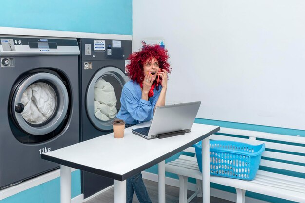 A smiling latin woman with red afro hair works with laptop and talks on the phone in laundry room