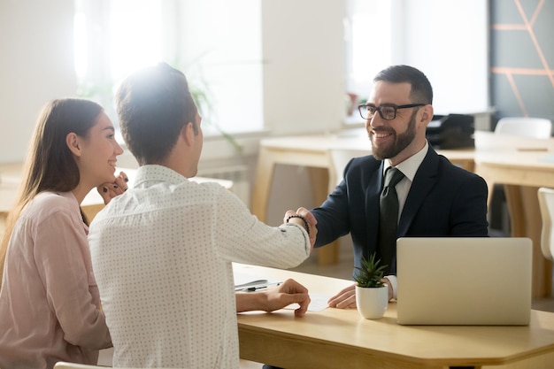 Smiling lawyer realtor or financial advisor handshaking young couple thanking for advice insurance broker or bank worker and millennial customers shake hands making deal investment or taking loan