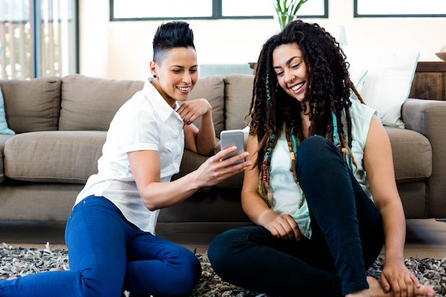 Smiling lesbian couple sitting on rug and looking at their mobile phone in living room