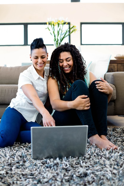 Smiling lesbian couple sitting on rug and using laptop in living room