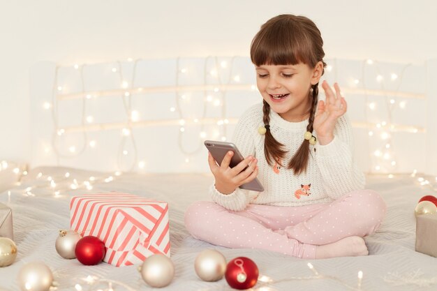 Smiling little adorable girl wearing white casual sweater sitting on bed with smart phone in hands, posing in festive bedroom, talking with friends or parents via video call, waving hand.