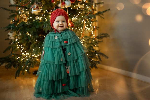 Photo smiling little baby girl in a christmas tree carnival costume and warm winter hat stands in front of a christmas tree new year celebration