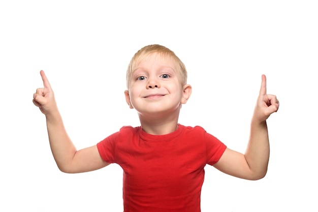 Smiling little blond boy in a red T-shirt is standing and pointing with his index fingers upwards