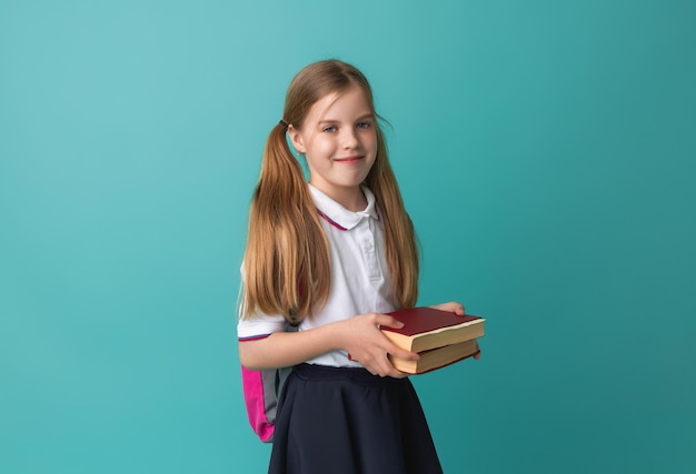 Smiling little blonde girl 1012 years old in school uniform with backpack holding books isolated on pastel blue background studio portrait The concept of children's lifestyle Education at school