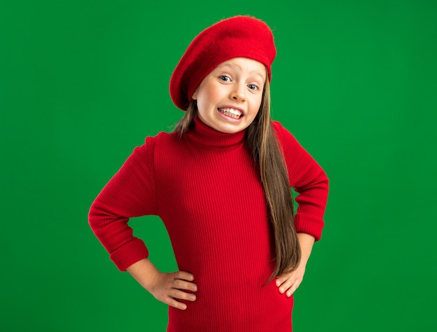 Smiling little blonde girl wearing red beret looking at front keeping hands on belly isolated on green wall with copy space