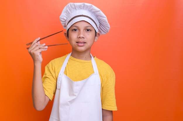 Smiling little boy in chef's toque and apron holding chopstick on orange background with copy space