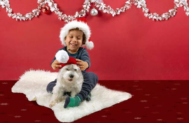 Smiling little boy playing with his puppy and Santa Claus hat in Christmas decorations