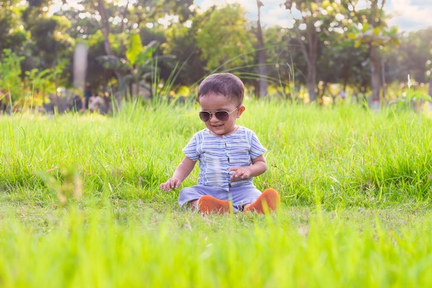 笑顔の小さな男の子が公園の芝生のフィールドに座って、サングラスをかけた男の子の赤ちゃん