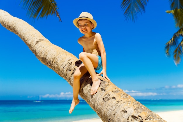 Smiling little boy in a straw hat having fun on a coconut tree on a sandy tropical beach. The concept of travel and family holidays.