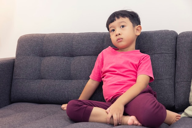 笑顔の小さな男の子がテレビを見て、リビングルームの快適なソファのリモコンを押す