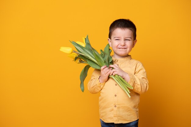 黄色のスタジオの背景に笑顔の小さな男の子。チューリップの花の花束と陽気な幸せな子。