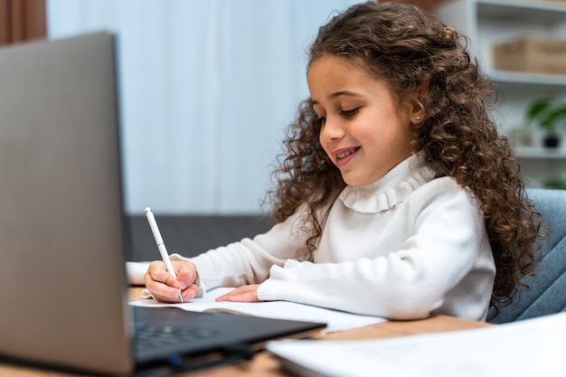 ラップトップを使用しながら注意深く何かを書いている小さな白人の女の子の笑顔コンピューターホームスクーリングの概念でオンラインで勉強する