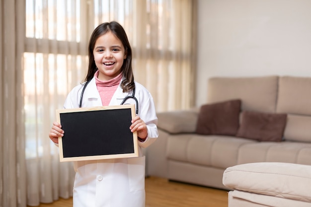 Piccolo bambino sorridente in uniforme del medico che tiene una lavagna a casa