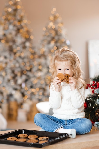 Smiling little child girl 34 year old wear knitted sweater and denim pants eating tasty baked biscuits on tray sit on table over Christmas tree and glowing lights Winter holiday season Good morning