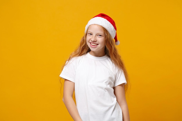 Smiling little ginger kid Santa girl 12-13 years old in white t-shirt, Christmas hat isolated on yellow background. Happy New Year 2020 celebration holiday concept. Mock up copy space. Looking camera.