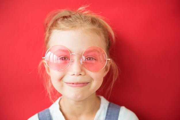 Smiling little girl in big round pink glasses on red