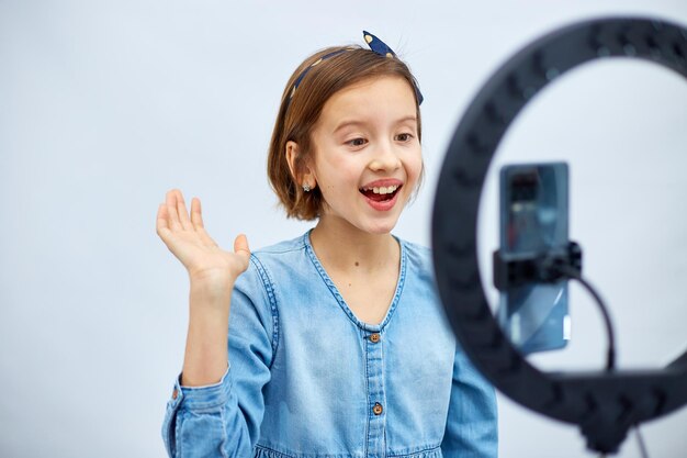 Smiling little girl blogger in casual denim dress use selfie led lamp and smartphone