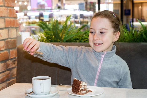 Smiling little girl in a cafe at the table pours sugar into a cup next to a plate with cake