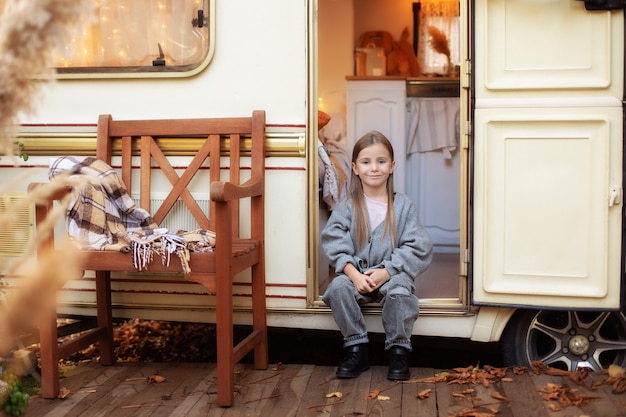 Smiling little girl in casual clothes sitting  near trailer door on porch RV house in garden