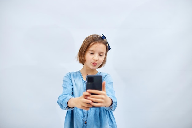 Smiling little girl in casual denim dress hold smartphone do selfie