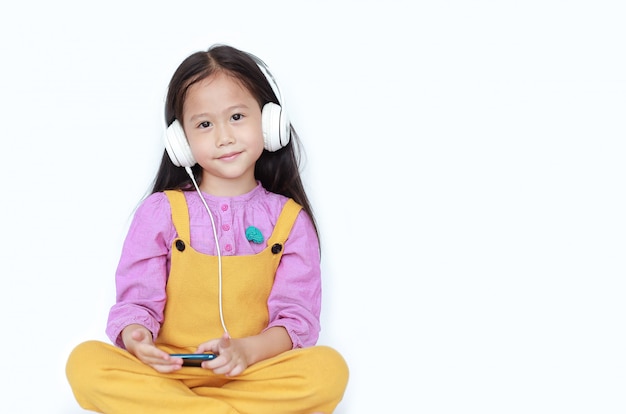 Smiling little girl enjoys listening to music with headphones isolated on white background with copy space.