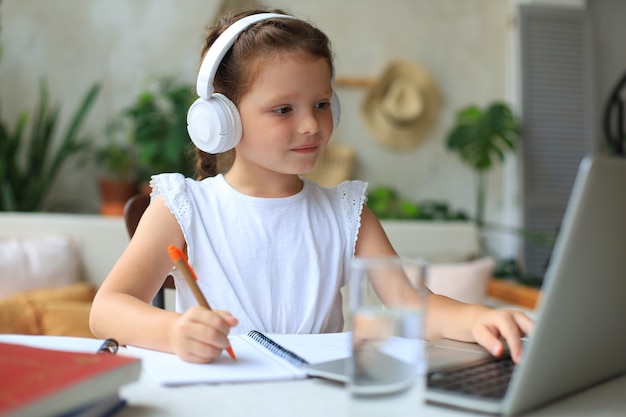 Smiling little girl in headphones handwrite study online using\
laptop at home, cute happy small child in earphones take internet\
web lesson or class on pc.