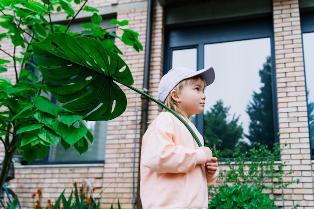 Smiling little girl holding monstera leaf in front of house in green garden