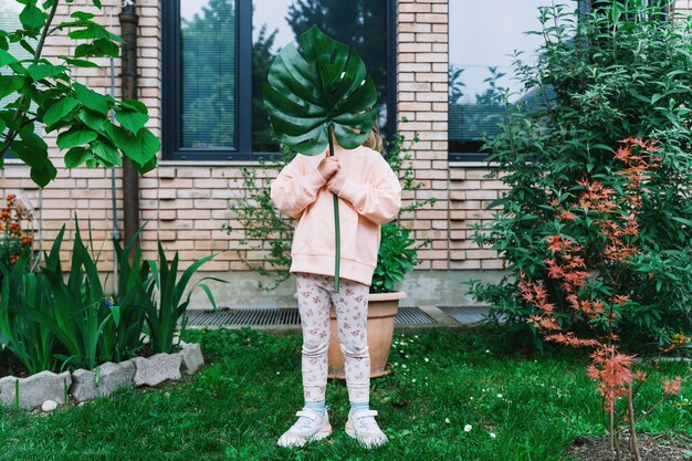 Smiling little girl holding monstera leaf in front of house in green garden