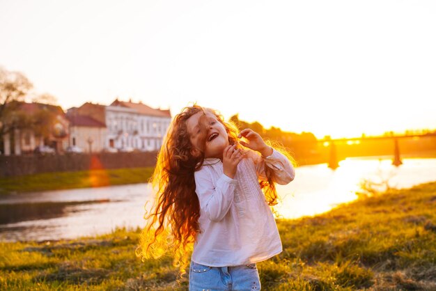 The smiling little girl is playing with her beautiful curly hair near the river