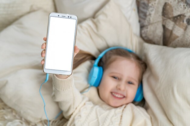 Smiling little girl kid in headphones shows white blank screen for text space