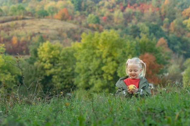 Фото Улыбающаяся маленькая девочка играет с яблоком в траве на фоне осеннего леса блондинка на пикнике на лугу