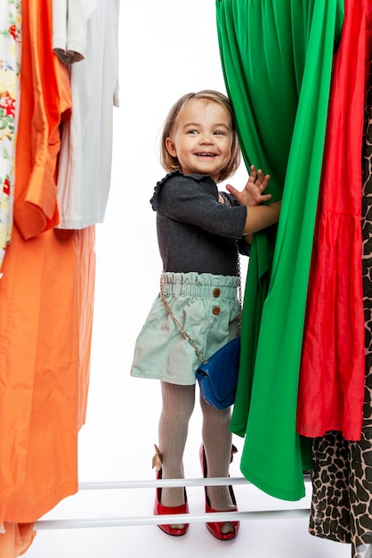 Smiling little girl in red shoes of mom among dresses on hangers. Delight, happiness and fashion. Vertical.