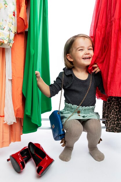 Smiling little girl in red shoes of mom among dresses on hangers. Delight, happiness and fashion. Vertical.