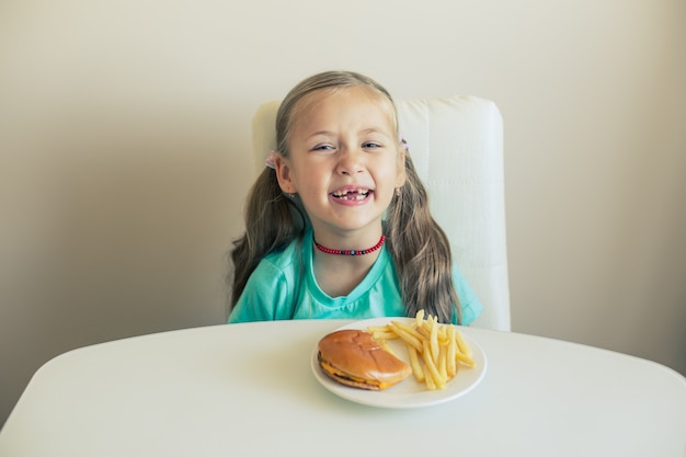 Улыбающаяся маленькая девочка сидит за столом с гамбургером и картофелем фри