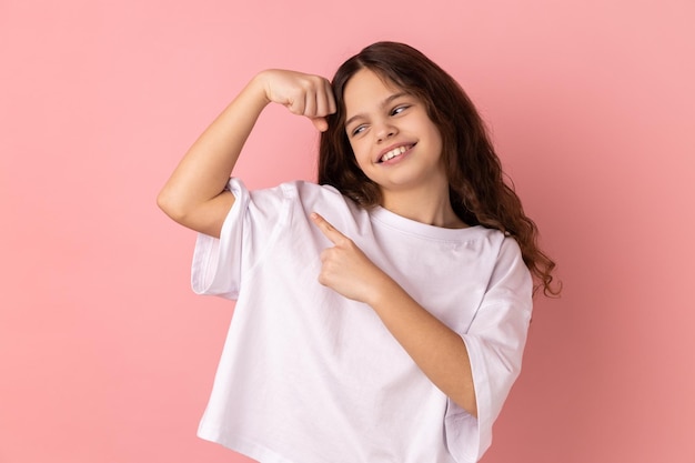 Smiling little girl showing her arm muscle and point it with finger proud of her strength