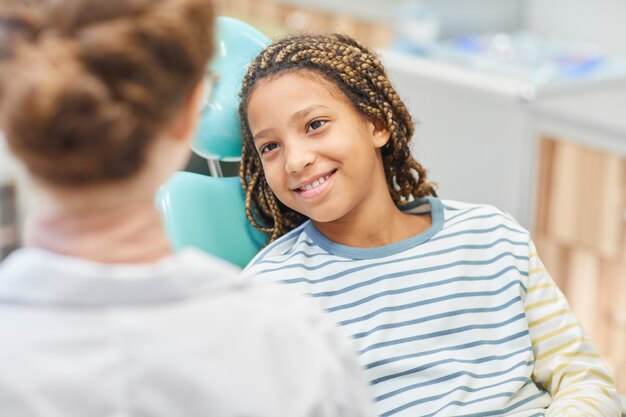 Photo smiling little girl sitting on dental chair and talking to doctor at hospital