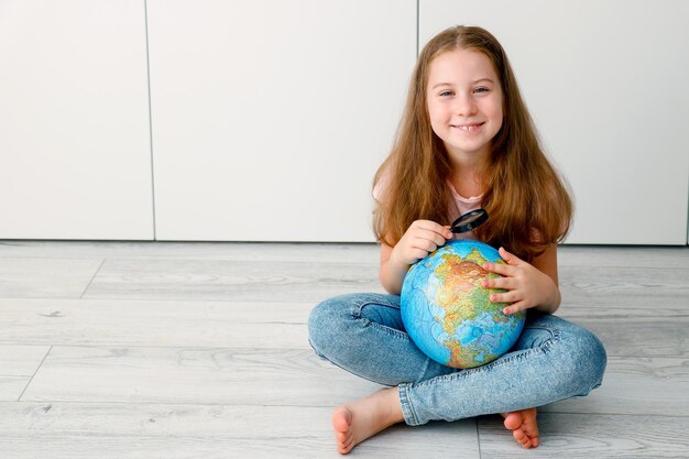 미소 짓는 작은 소녀가 지구와 확대 유리창으로 바닥에 앉아 있습니다.