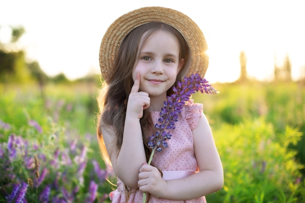 Smiling little girl in straw hat holds lupine flower in her hands at a field